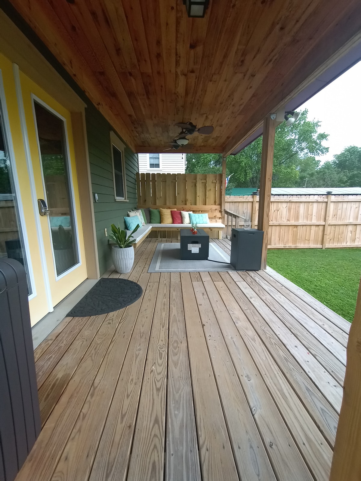 A covered porch is shown with wooden flooring and a slatted ceiling. A seating area is decorated with colorful cushions, positioned alongside a potted plant. A small table sits nearby, complementing the outdoor space which is enclosed by a wooden fence.