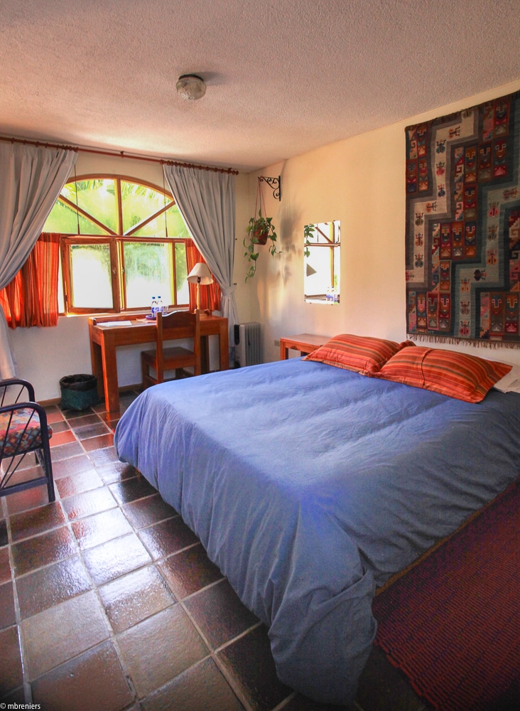 A well-lit room features a spacious bed with light blue bedding, complemented by striped accent pillows. A wooden desk is positioned by a large window, adorned with orange curtains. Decorative wall art and tile flooring enhance the room's simple yet comfortable design.