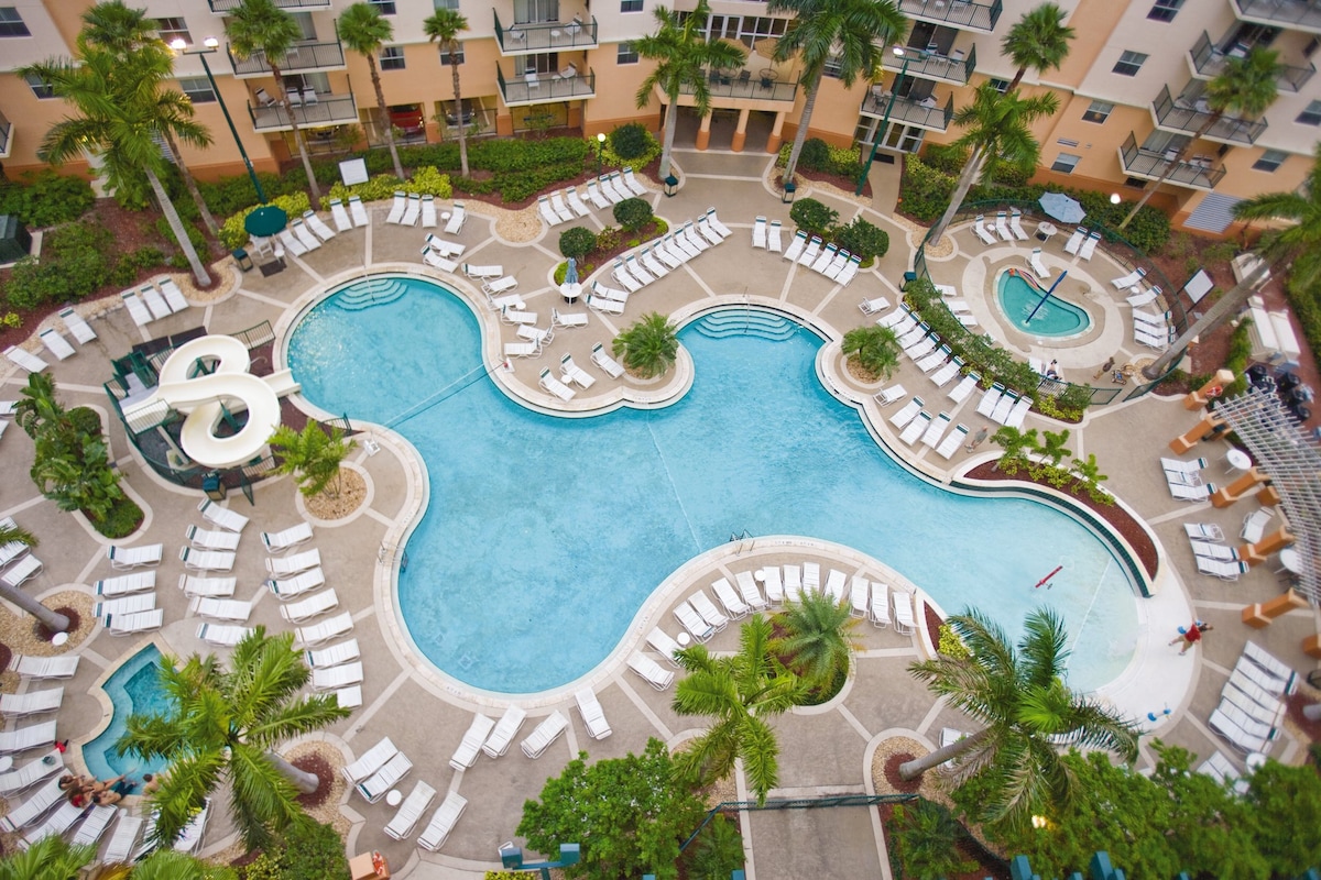 An aerial view captures a resort pool shaped in an intricate design, surrounded by lounge chairs and tropical landscaping. Two hot tubs are visible next to the main pool, and palm trees provide shade throughout the area, creating a refreshing oasis.