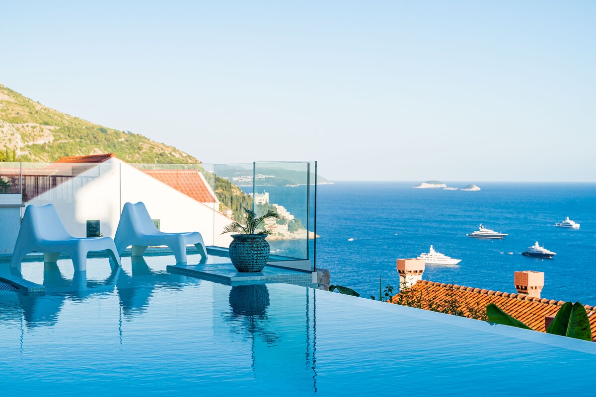 An infinity pool is shown with calm water reflecting the sky, surrounded by a glass railing. Two sleek white lounge chairs are positioned at the edge, overlooking a scenic view of the Adriatic Sea and distant boats. Lush greenery is visible along the pool's edge.