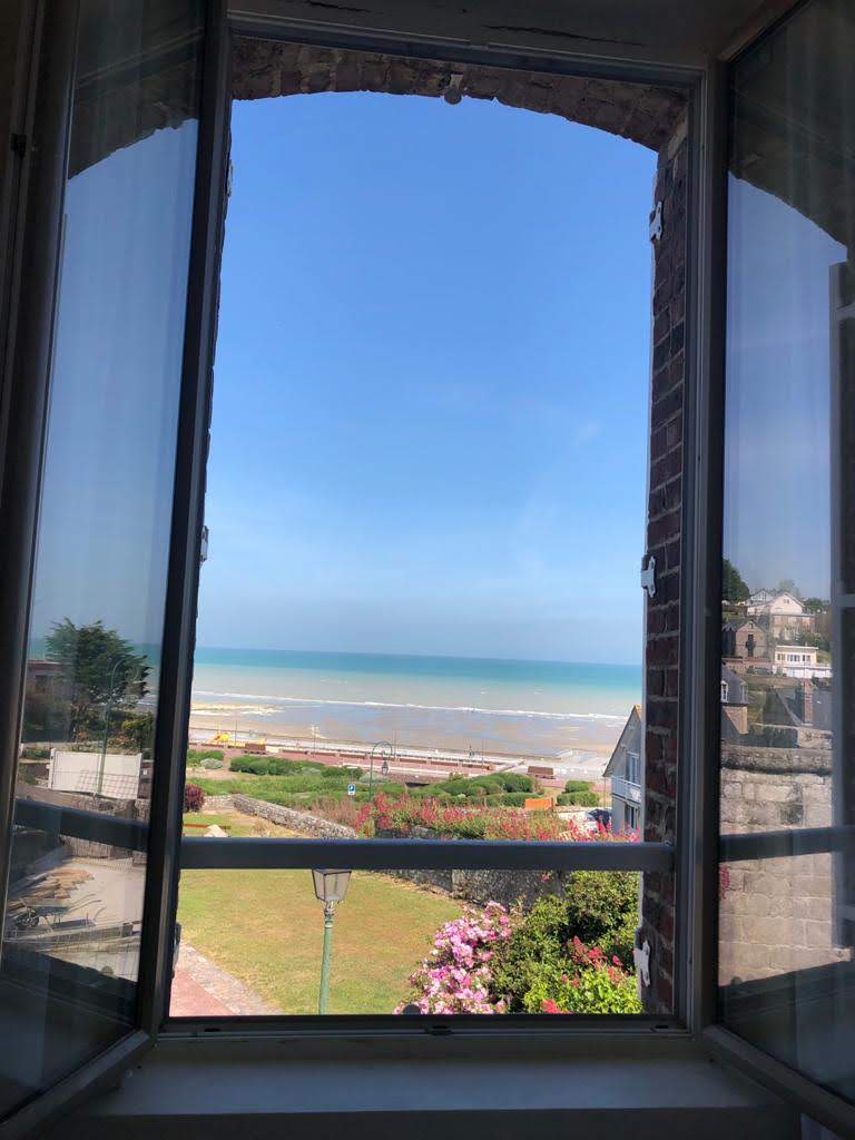 A view is captured through open windows, revealing a panoramic scene of the sea and a lush garden below. Vibrant flowers line the pathway, and the shoreline is visible against a clear blue sky, enhancing the connection to the outdoor environment.