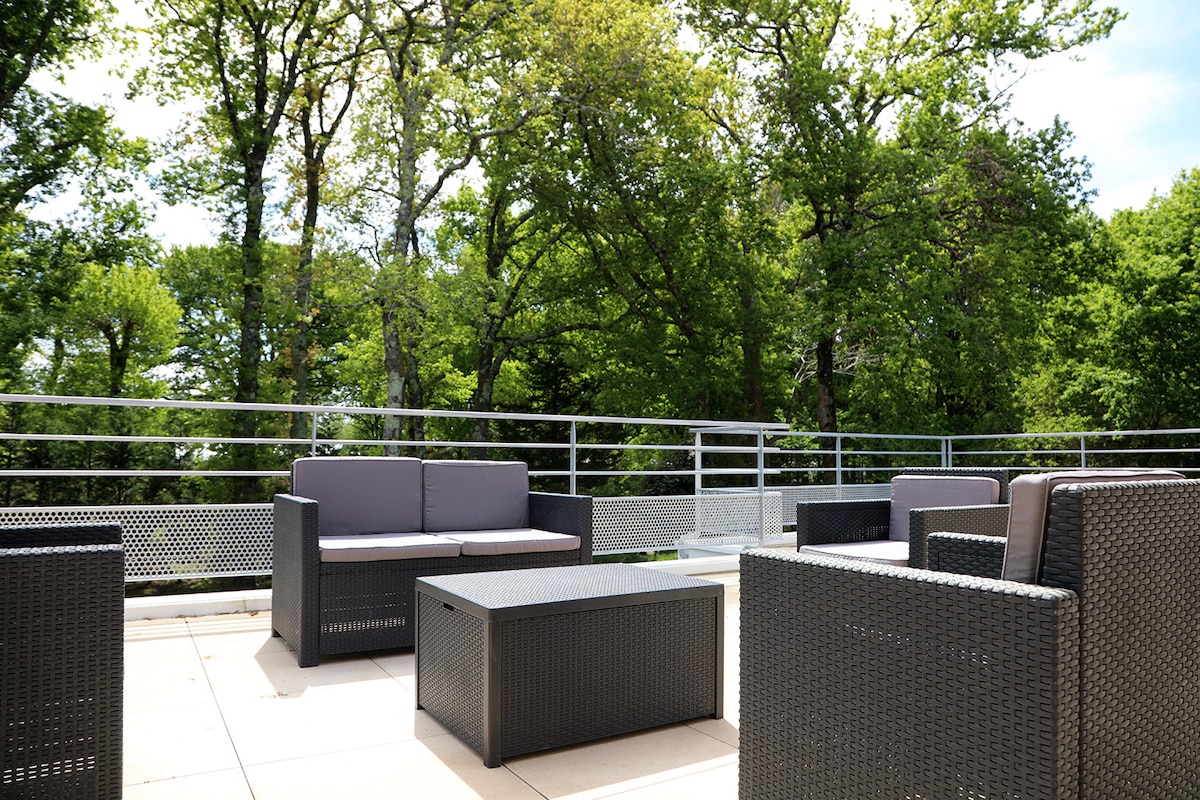 A private terrace features modern outdoor furniture, including several woven seating arrangements and a coffee table. Lush greenery from surrounding trees creates a peaceful backdrop, while a railing provides a sense of openness to the space.