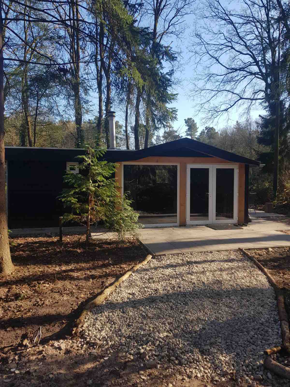 A chalet is situated amidst trees, featuring a sloped roof and large glass doors at the entrance. A pathway of gravel leads to the door, surrounded by natural landscaping and greenery.