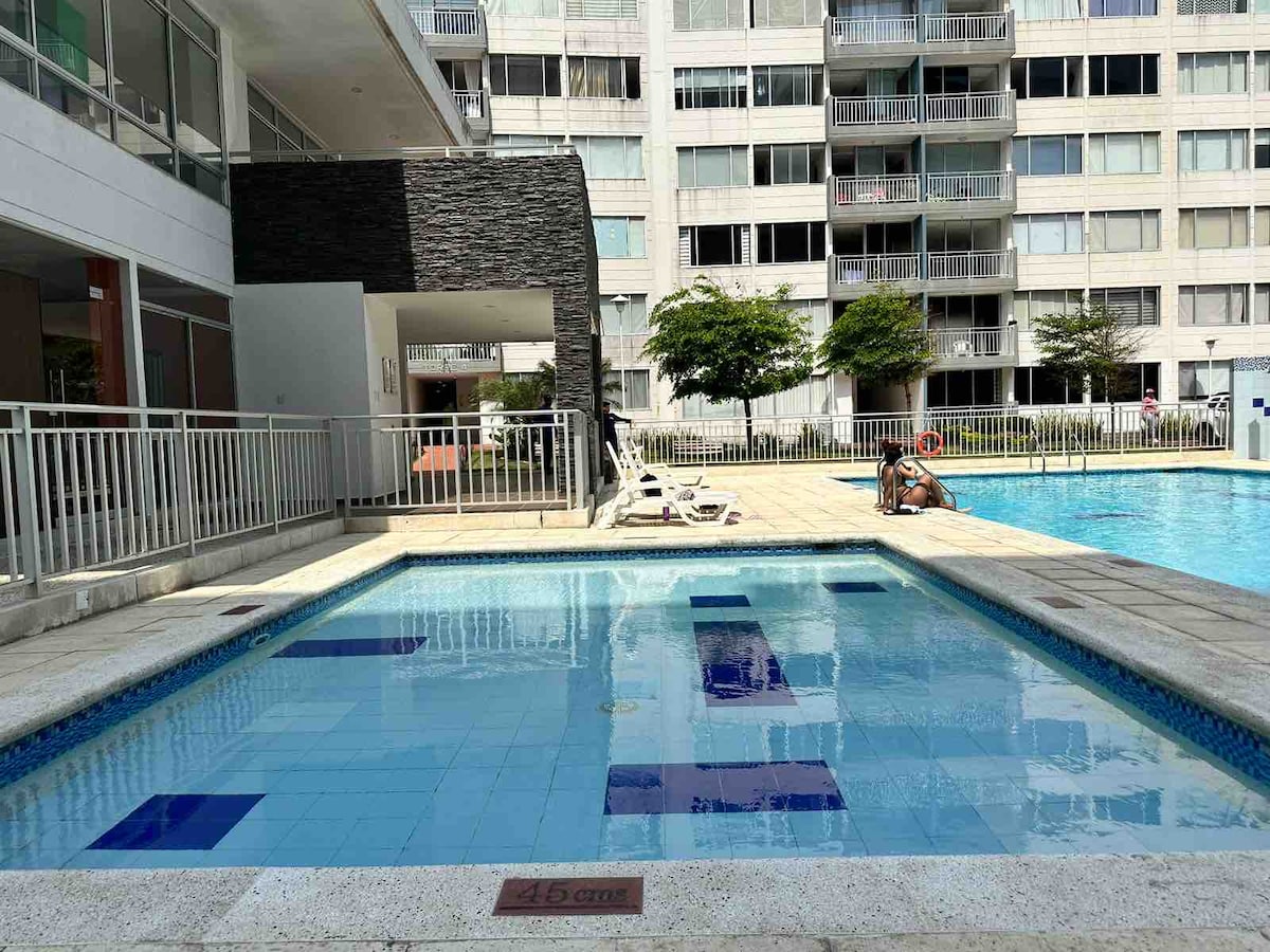 Two swimming pools are visible, including a smaller shallow area with a depth marker of 1.5 meters. Sun loungers are positioned around the pool area, and landscaping adds a touch of greenery to the surrounding space. The buildings in the background feature balconies and large windows.