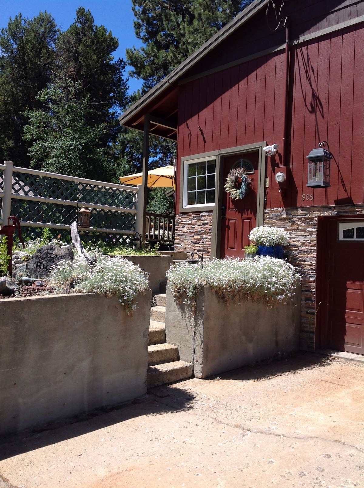 The exterior features a vibrant red house with stone accents, surrounded by flowering plants in concrete planters. Steps lead up to a welcoming entryway, while a shaded patio is visible above, framed by a decorative fence and lush trees in the background.