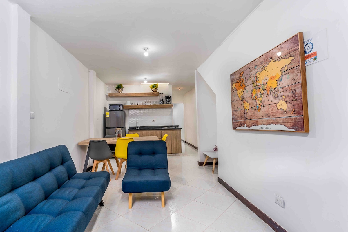 A modern living area features a blue sofa and a cozy chair, arranged around a wooden coffee table. A dining area with yellow and wooden chairs is visible, along with a fully equipped kitchen in the background. A world map is displayed on the wall.