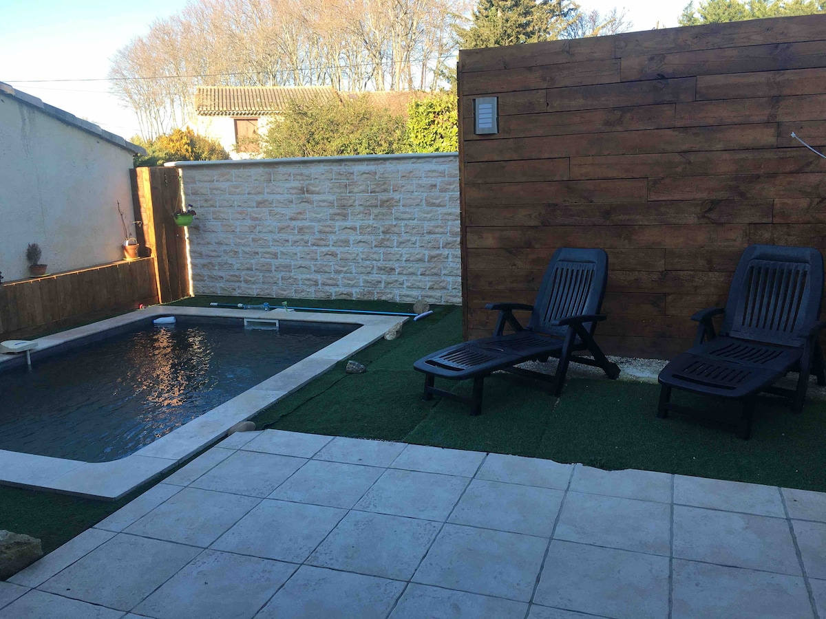 A private outdoor space is presented, featuring a shallow rectangular pool bordered by stone. Two black lounge chairs are positioned on artificial grass, complementing a textured wooden wall behind them. Sunlight highlights the clean tile flooring, contributing to the serene setting.