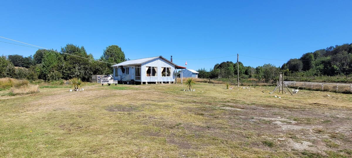 A small vacation house is shown in a spacious, open landscape, surrounded by greenery. The structure features large windows and a covered porch, inviting natural light. The surrounding yard is mostly grass, with a few trees and a distant view of hills under a clear blue sky.