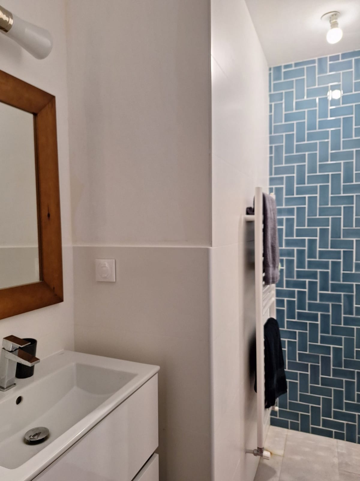 A modern bathroom is displayed, featuring a compact sink with a mirror framed in wood. A shower area is visible, showcasing blue tiles with a herringbone pattern. Towel racks hold neatly arranged towels, and bright overhead lighting enhances the clean and functional space.