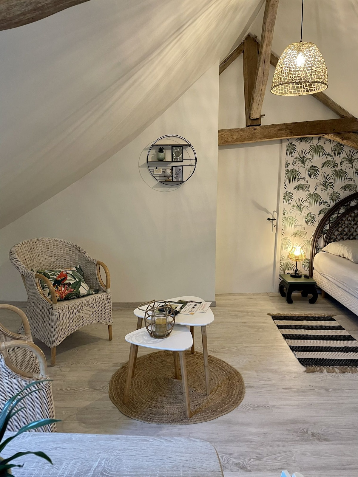 A cozy seating area features two woven armchairs beside a circular jute rug. A small table with decorative items is positioned between the chairs. The room showcases wooden beams and is accented by a light fixture made of natural materials, creating a serene ambiance.