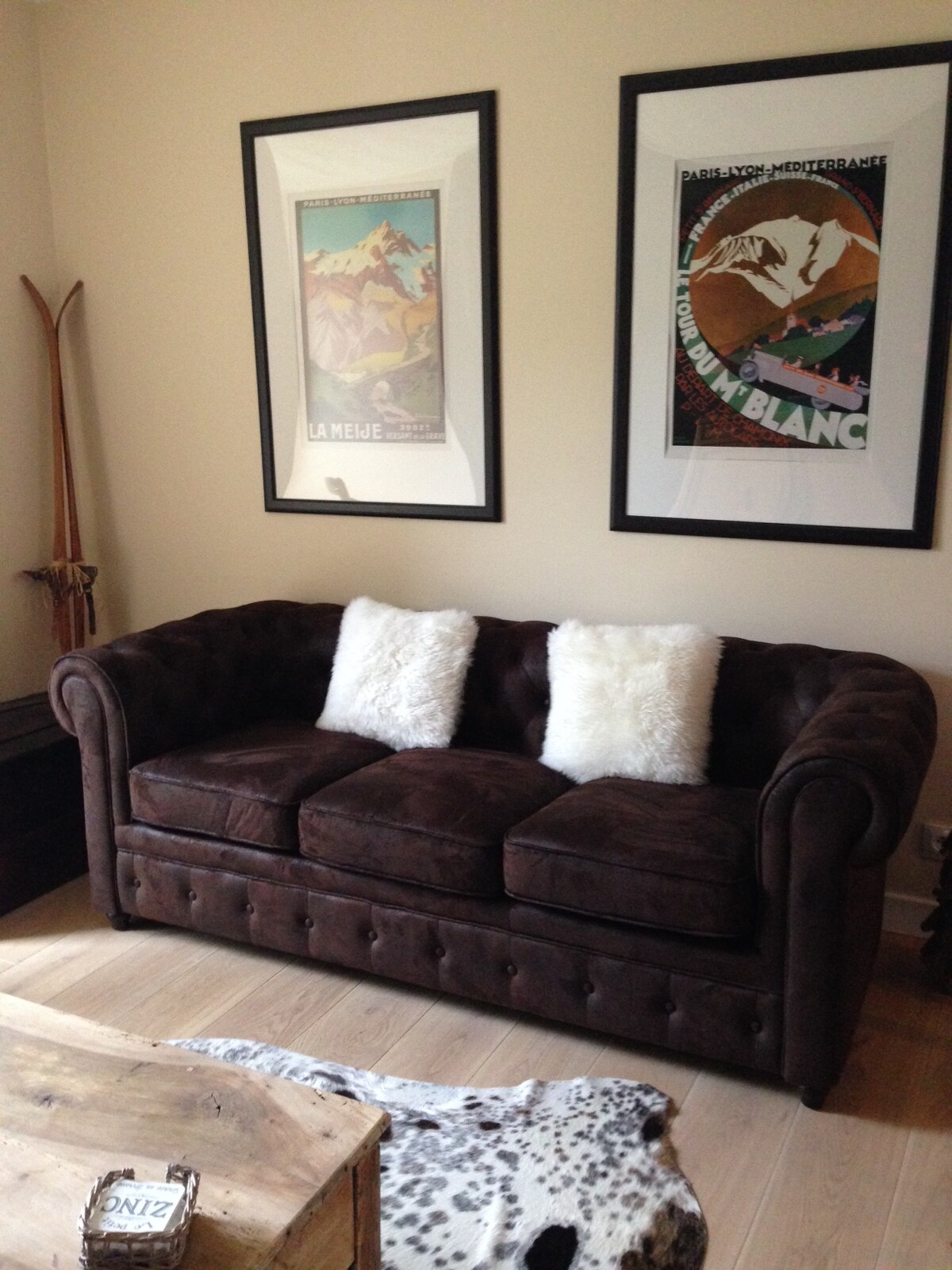 A welcoming living area is highlighted by a dark brown tufted sofa adorned with two white fur pillows. The walls display framed vintage posters, while a wooden coffee table and a cowhide rug complete the cozy atmosphere.