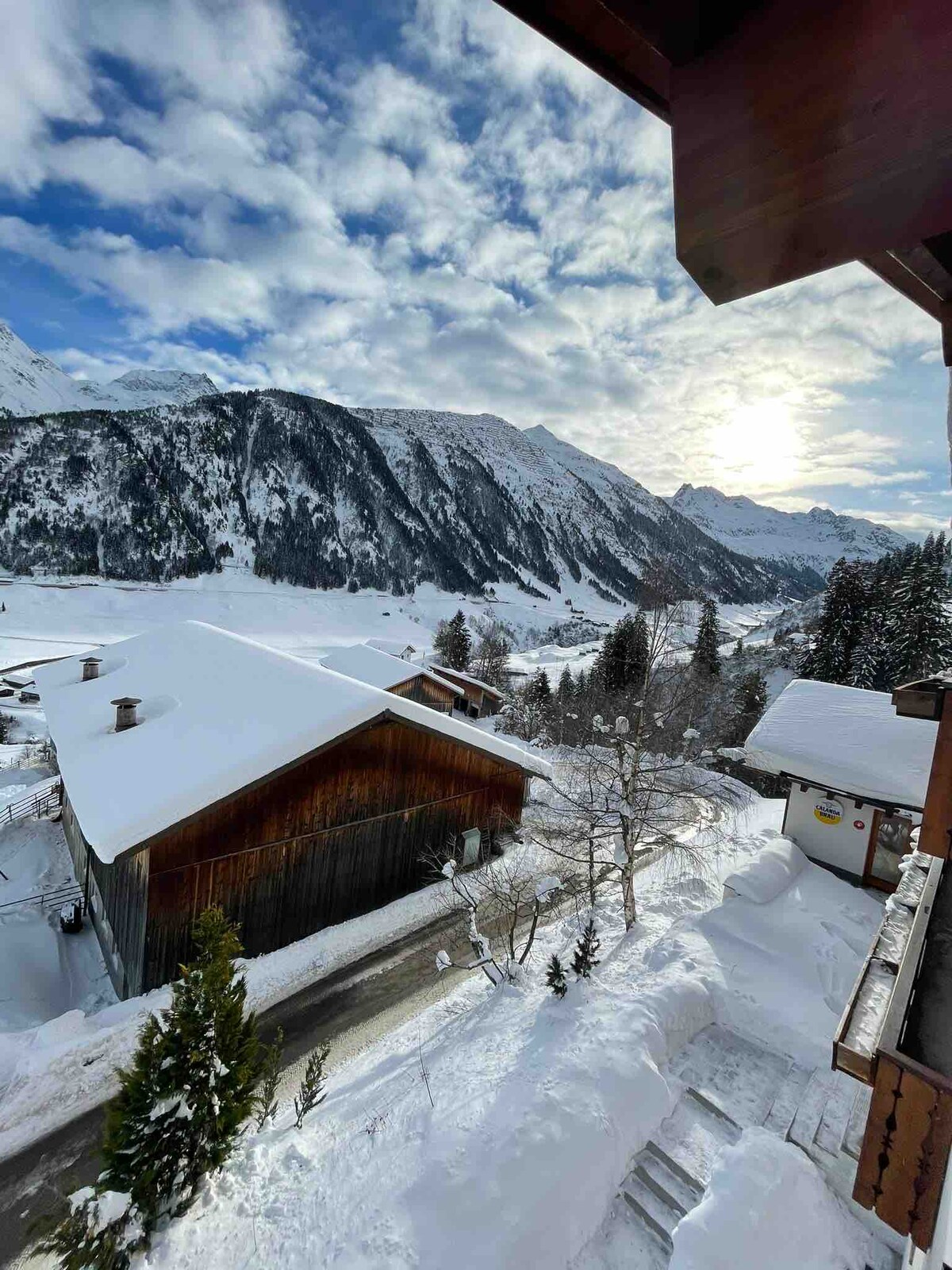 A winter landscape showcases snow-covered mountains under a partly cloudy sky, with sunlight breaking through. In the foreground, a wooden building is visible, complemented by snow-laden trees and a winding path. The tranquil scene conveys a sense of mountain serenity.