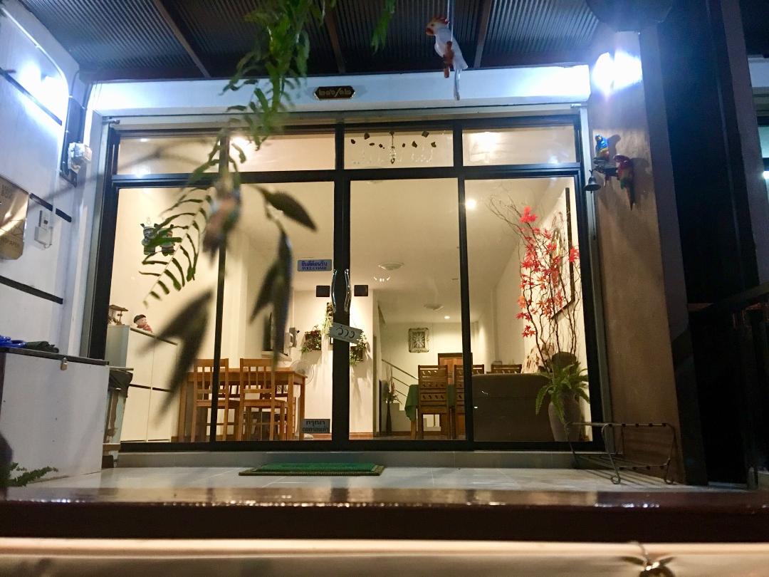 The entrance features large glass doors that provide a view into the open dining area. A clean, welcoming space is visible, decorated with plants and minimalistic furniture. Soft lighting adds to the inviting atmosphere, while a doormat sits on the threshold.