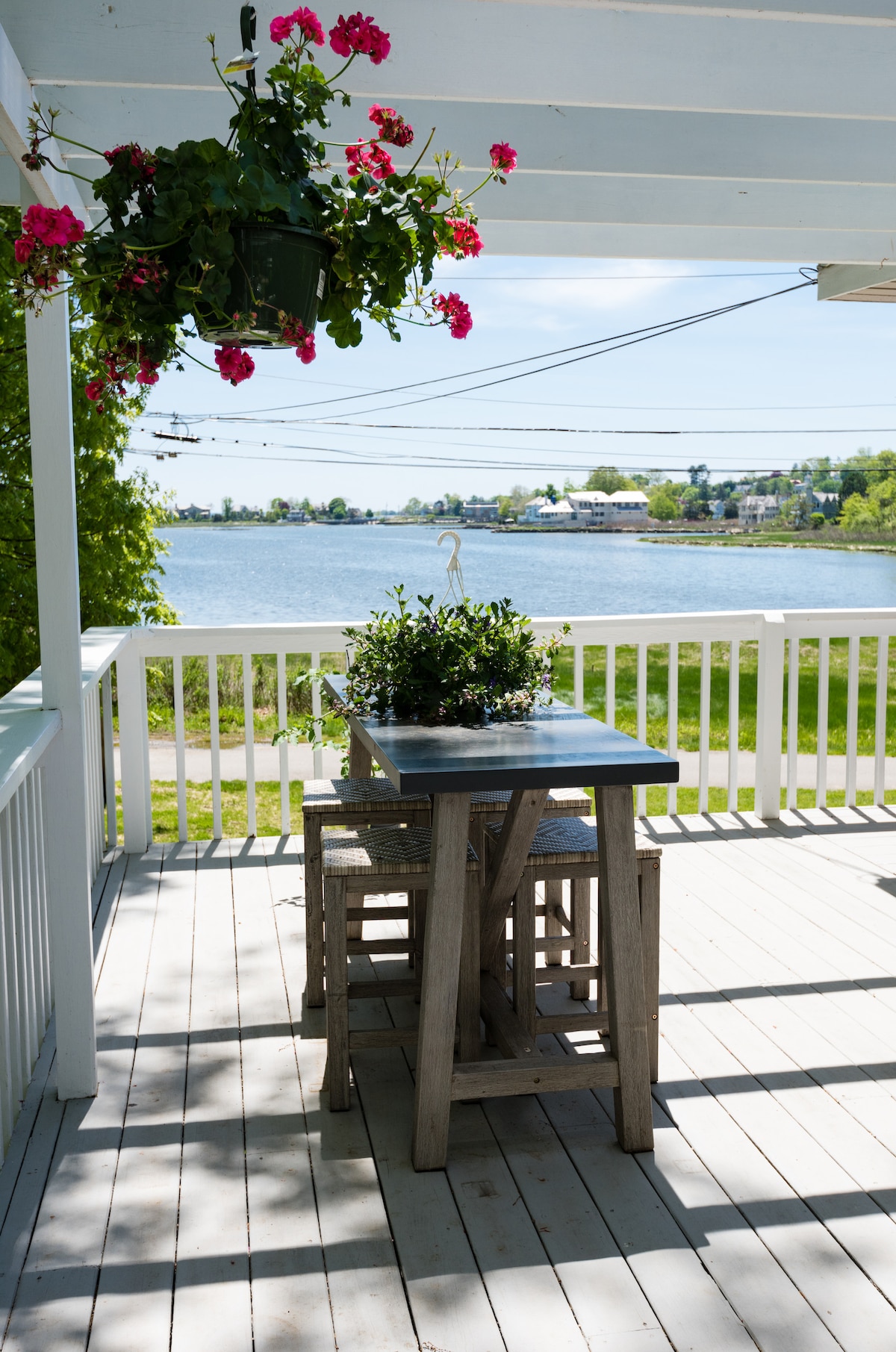 An outdoor deck area features a small, weathered wooden table surrounded by tall stools. Greenery and vibrant pink flowers drape from a hanging planter above, while a scenic view of Mill Pond is visible in the background, highlighting the tranquil water and surrounding nature.