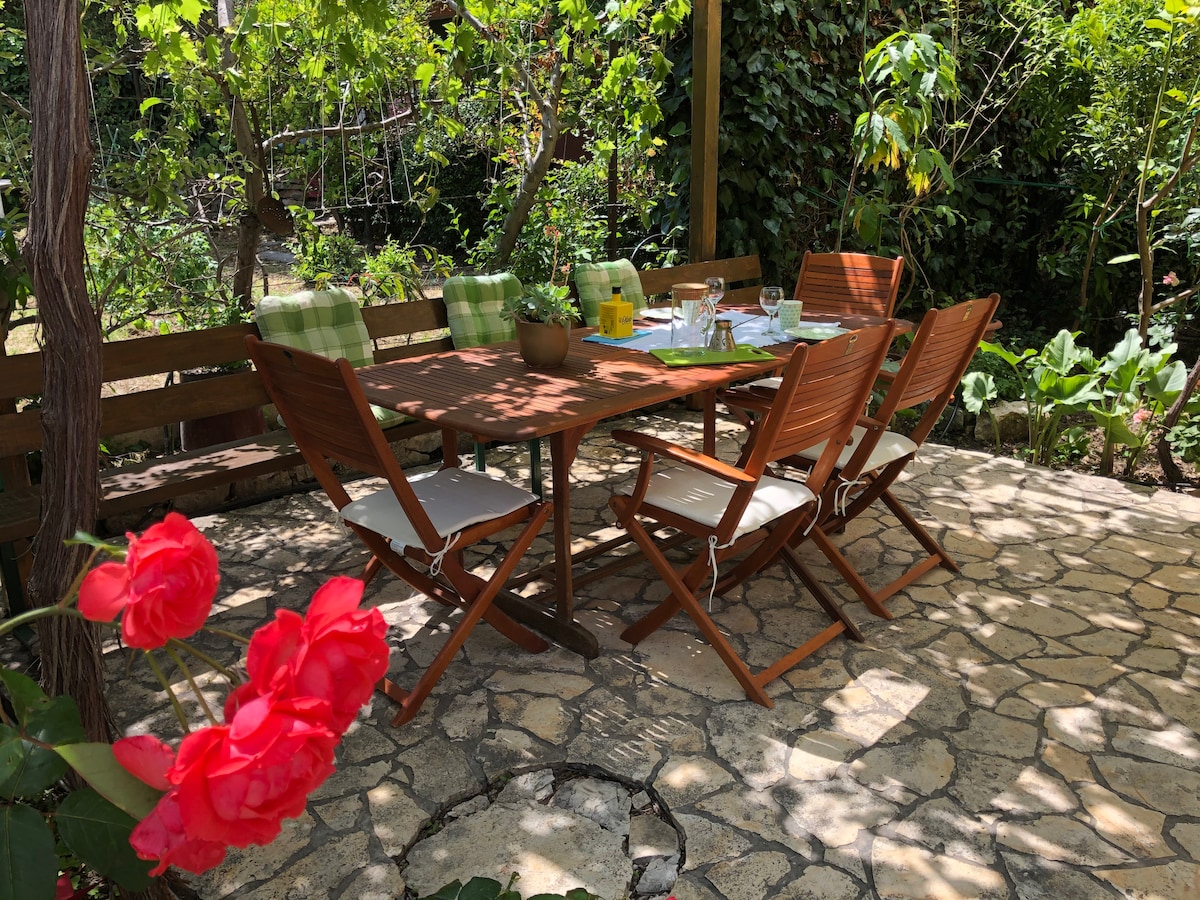 A dining area is positioned among lush greenery, featuring a wooden table surrounded by six folding chairs. Sunlight filters through the leaves, creating dappled patterns on the stone floor. A potted plant and glasses are arranged on the table, evoking a pleasant atmosphere for outdoor dining.
