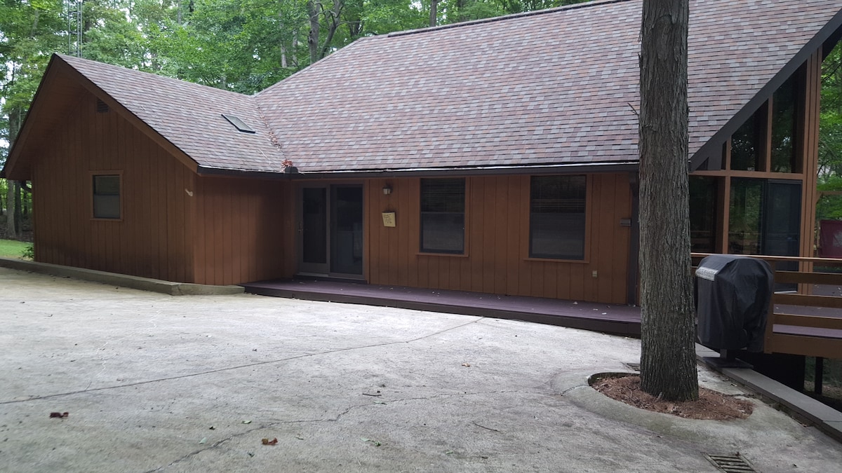A modern cabin exterior is seen, showcasing a combination of wood paneling and large glass windows. A spacious driveway leads to the entrance, which is bordered by a gravel area. A gas grille is positioned on the side, surrounded by lush greenery.