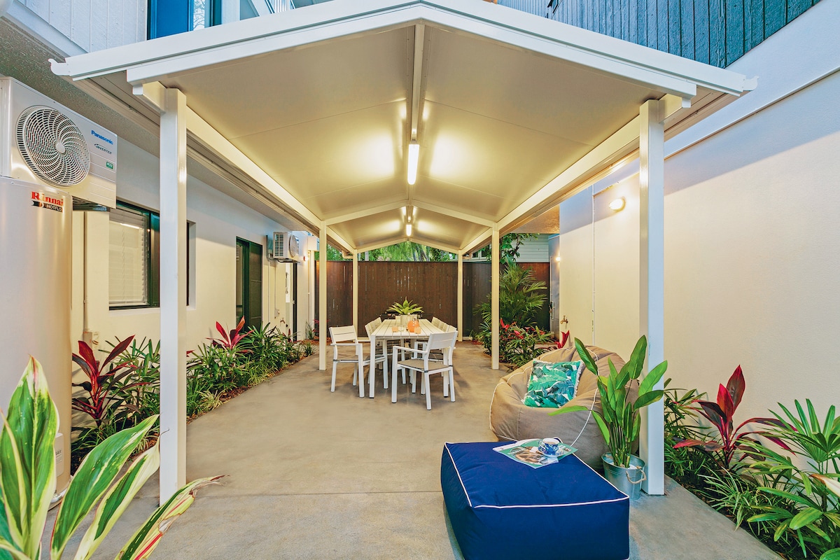 An outdoor patio area is covered with a roof, providing a sheltered space for relaxation. A dining table with several chairs is visible, surrounded by lush greenery and decorative plants. A seating area is comprised of a cozy bean bag and a cushion, inviting leisure.