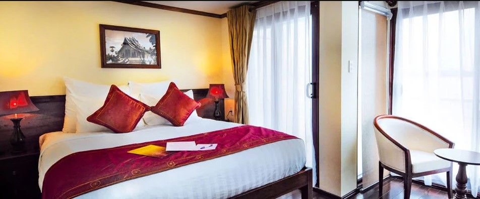 A modern bedroom features a spacious bed adorned with decorative cushions and a vibrant throw. Soft lighting from bedside lamps complements natural light filtering through sheer curtains. A small table and chair are positioned nearby, creating a cozy reading nook.