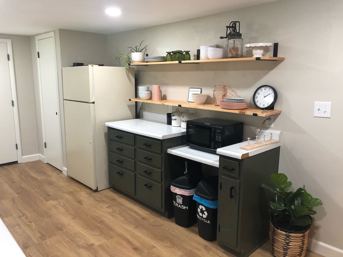 A well-equipped kitchen area features green cabinets and a white countertop. Open shelving displays various dishware and decorative items, while a clock and a microwave are positioned on the counter. A refrigerator stands at the end, and bins for trash and recycling are neatly organized below.