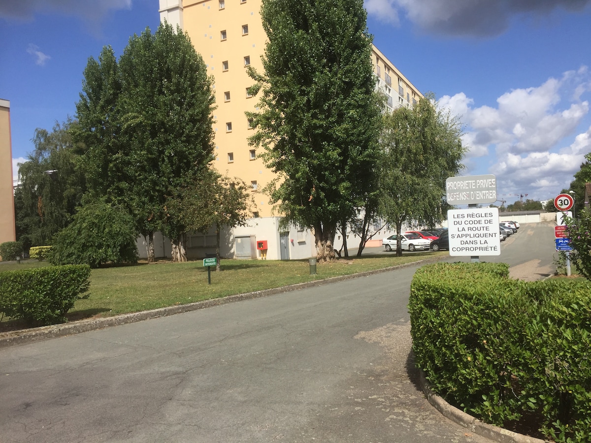 A serene view of the apartment building surrounded by well-maintained green lawns and trees. The inviting path leads towards the entrance, with designated parking visible in the background. A sign offers information about the property, enhancing the tranquil environment.