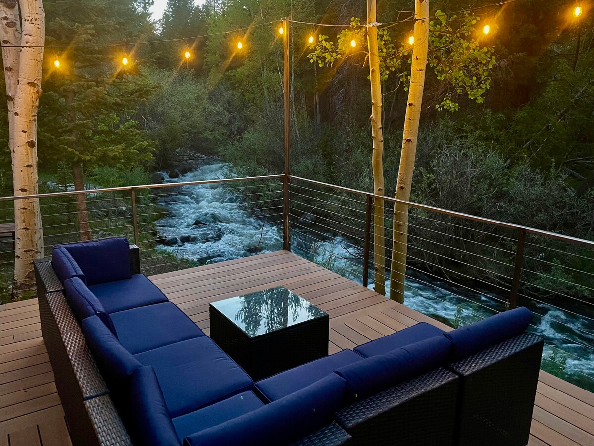 A spacious outdoor deck features a comfortable seating arrangement with dark wicker furniture and a glass-topped table. String lights illuminate the area, and a view of flowing water can be seen just beyond the railing, framed by lush greenery and trees.