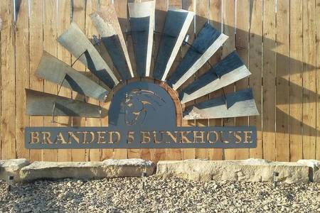 A decorative sign reading 'Branded 5 Bunkhouse' is mounted on a wooden fence. The background features a stylized sun design created with metal blades, positioned above the sign, lending a rustic charm to the property.