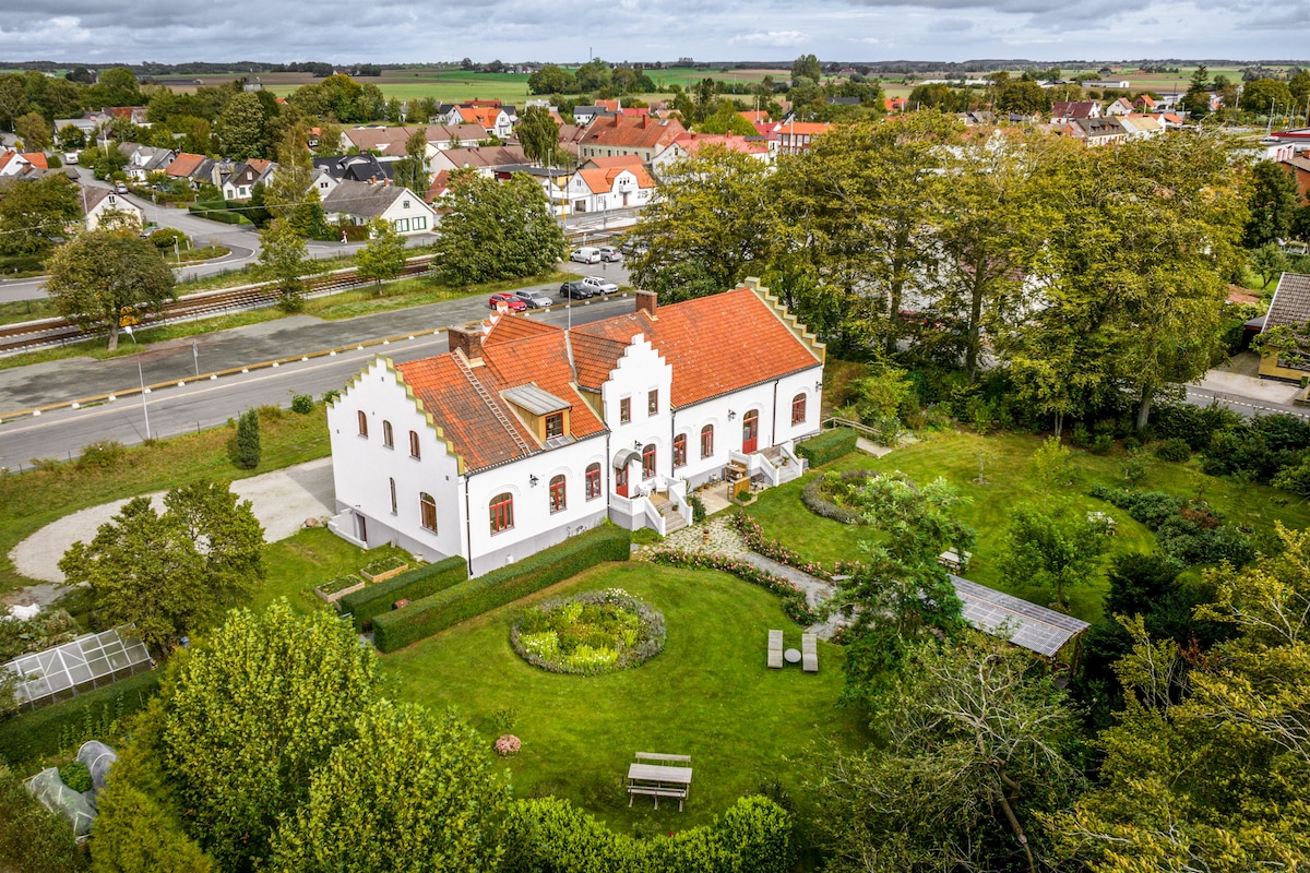 The property features a charming white house with a red-tiled roof, surrounded by a landscaped yard. Scenic greenery and garden areas are visible, along with several seating spaces. The neighborhood is characterized by suburban homes and open fields in the background.