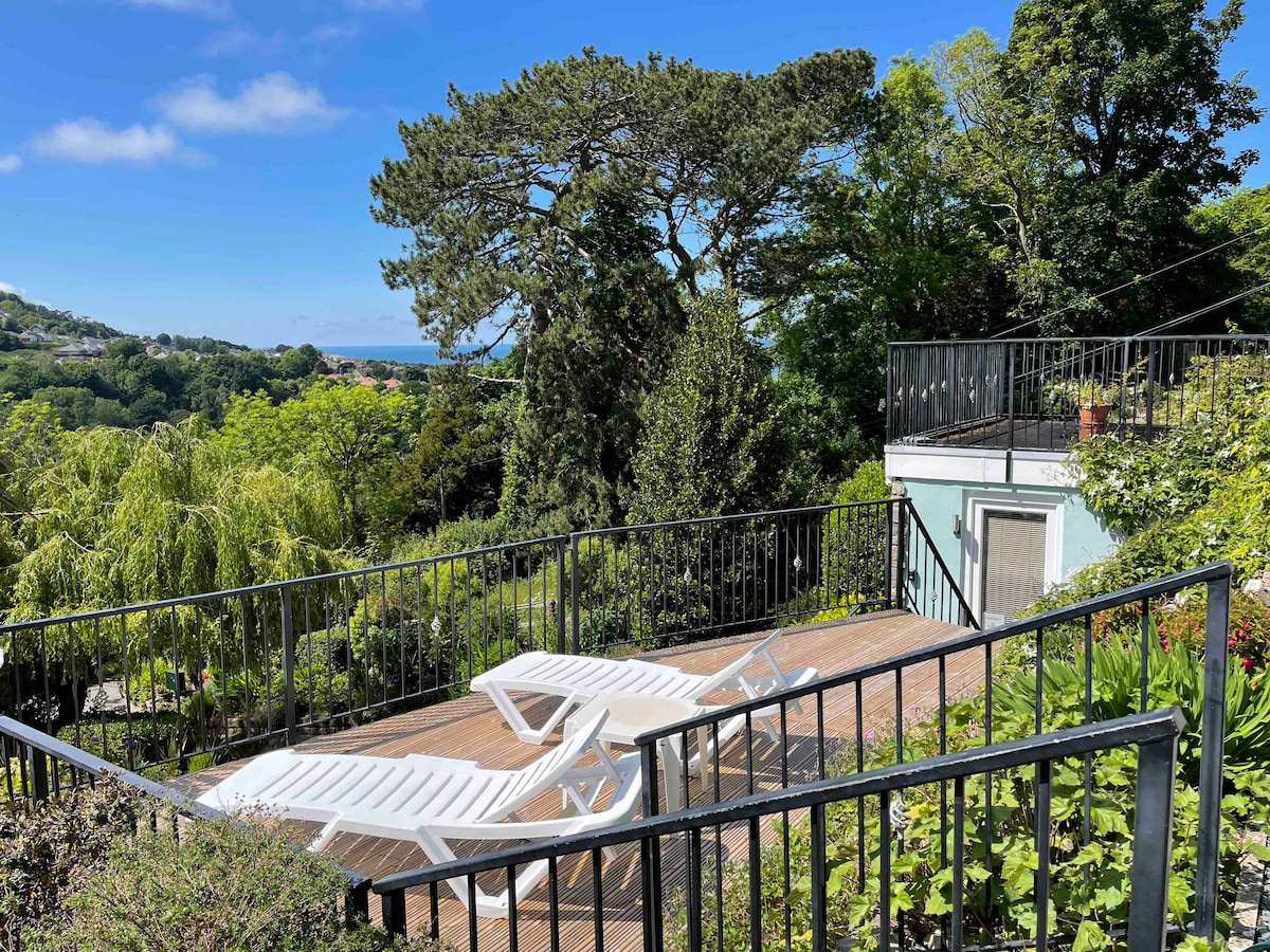 A spacious terrace surrounded by lush greenery offers views of the elevated landscape. Sun loungers are arranged on the wooden decking, with a railing providing safety. Fresh foliage and trees contribute to a tranquil outdoor environment.