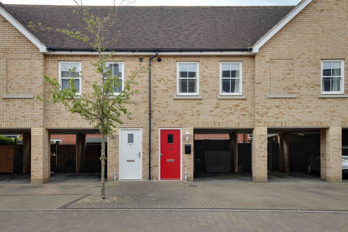 A charming brick exterior presents two connected homes, distinguished by a vibrant red door on one and a white door on the other. Parking spaces are provided underneath the structure, with a tree adding a touch of greenery to the well-maintained walkway.