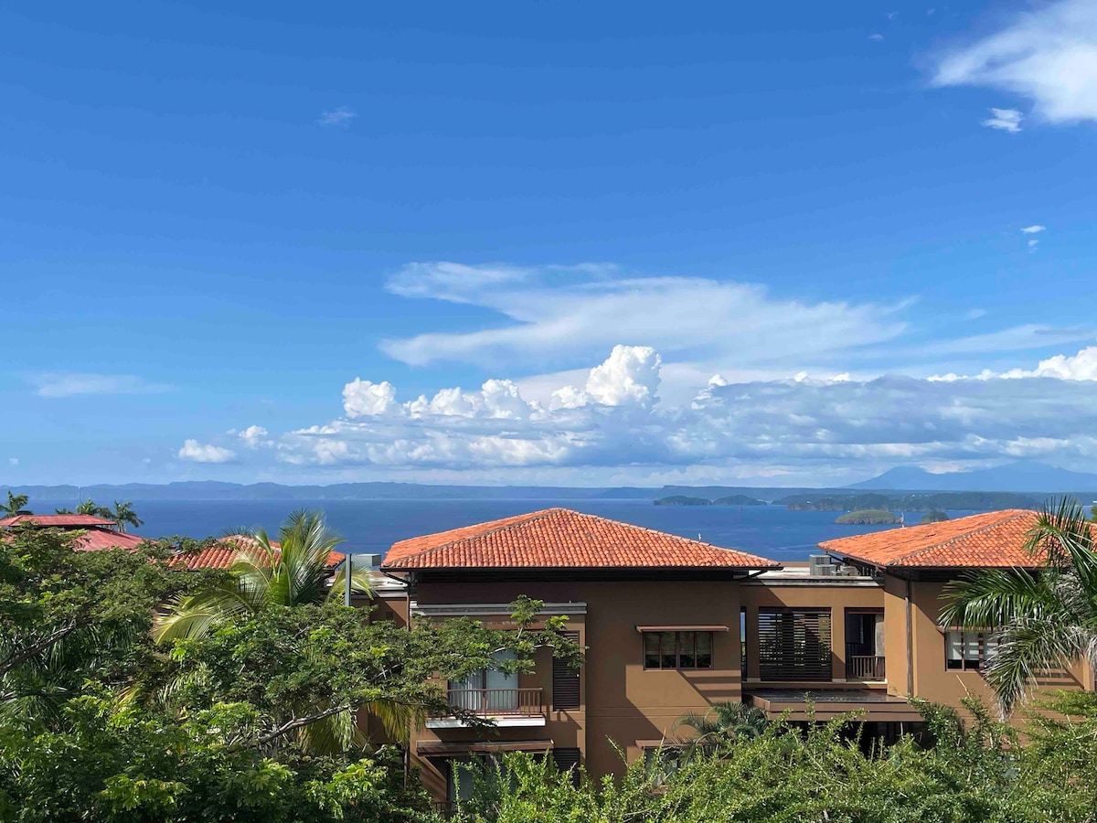 A panoramic ocean view is presented, showcasing the vibrant blue waters and distant islands under a clear sky. The scene includes terracotta rooftops of nearby buildings framed by lush greenery, emphasizing the tranquil setting in a hilly area.