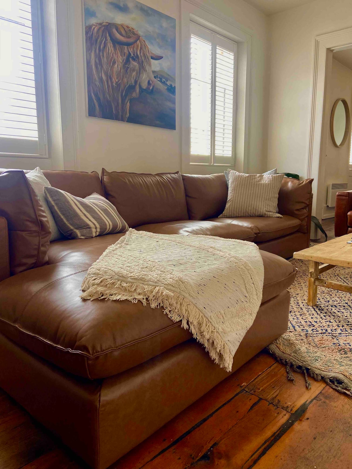 A spacious brown leather sofa is positioned in a well-lit living area. Decorative pillows are arranged on the sofa, accompanied by a textured throw blanket. Natural light filters through the plantation shutters, highlighting the wooden floor beneath. A round mirror and a coffee table are visible in the background.