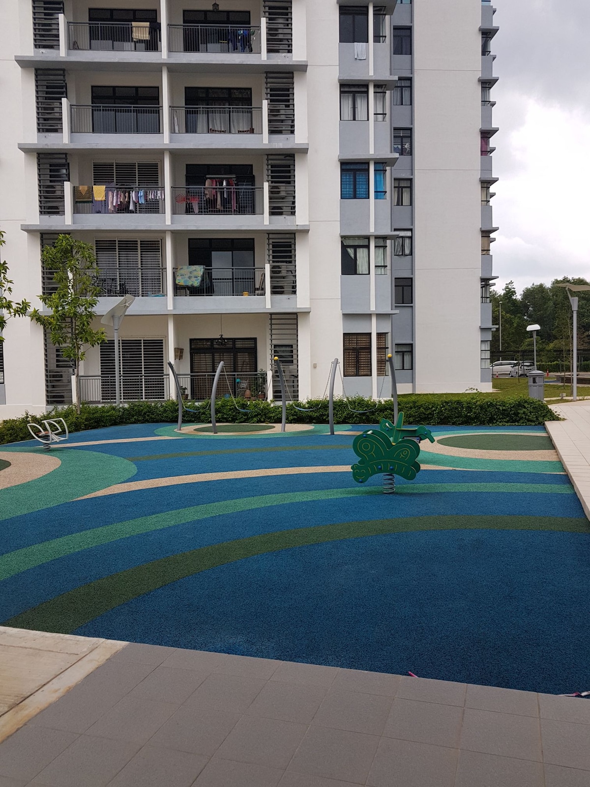 A playground area features colorful, patterned flooring in shades of blue and green, with two swings positioned on a soft, safe surface. Surrounding greenery enhances the outdoor space, with the building façade visible in the background, showcasing multiple windows and balconies.