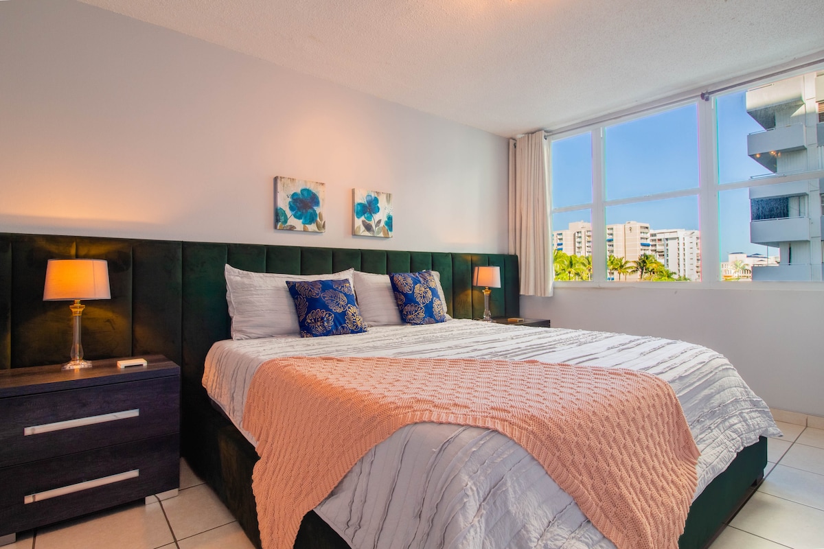 A spacious bedroom features a large dark bed covered with a striped blanket. Two blue floral art pieces are mounted above the bed, and bedside tables with lamps flank either side. Large windows allow natural light to fill the room.