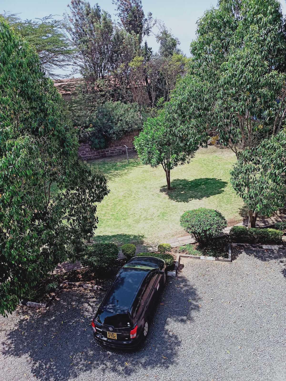 A gravel parking area is visible, accommodating a black vehicle. Lush greenery surrounds the area, including tall trees and manicured bushes, leading to a well-maintained lawn in the background. The scene reflects an outdoor space that offers a sense of tranquility.