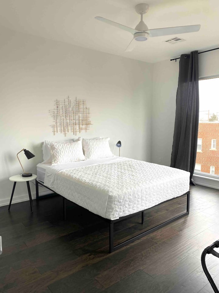 Minimalist bedroom with black metal bed, white textured bedding, and geometric wall art. Dark wood floors and natural light from black-curtained windows create a serene, modern retreat.