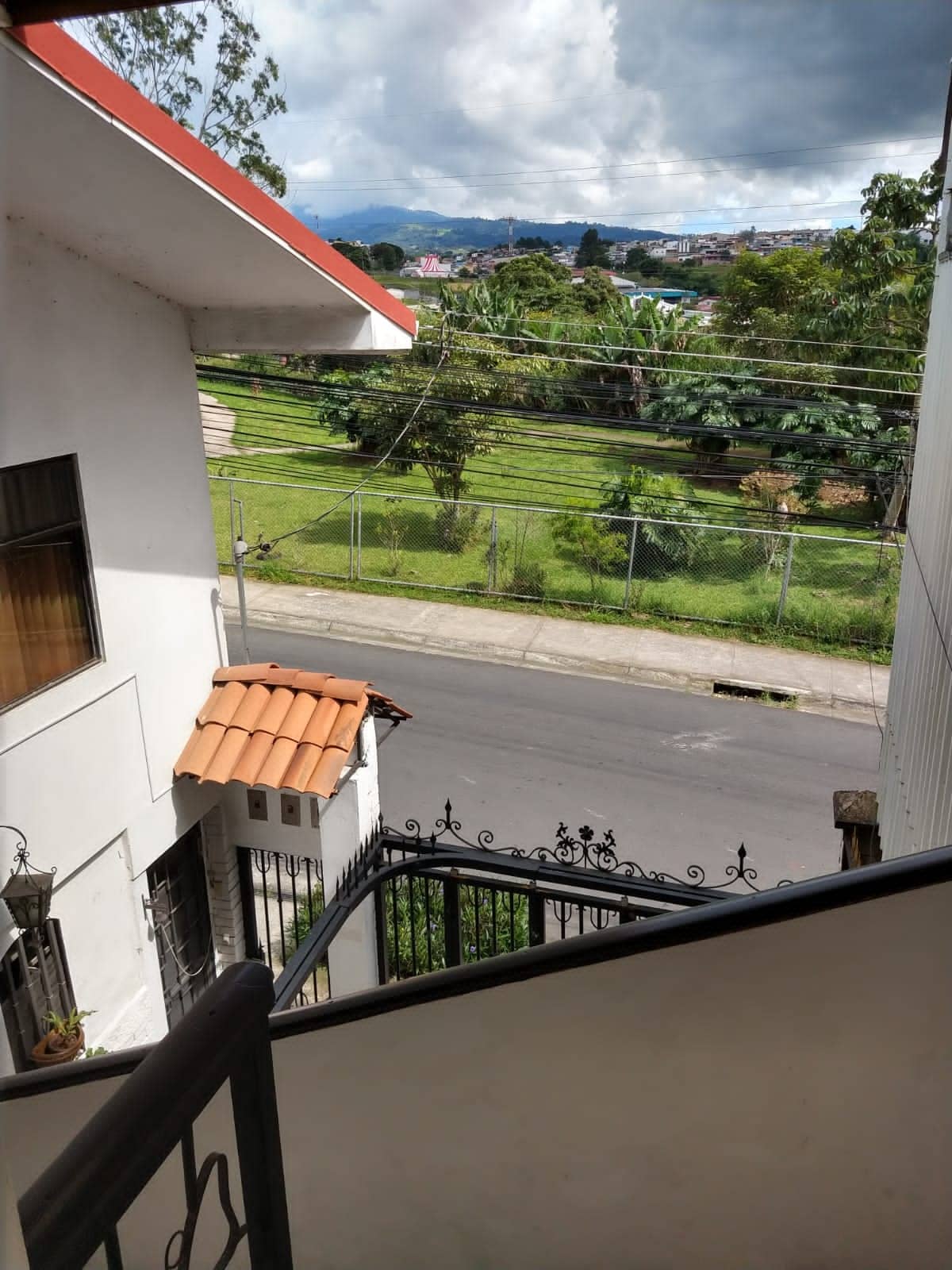 An exterior view from a balcony showcases a landscaped green area with trees and plants. A winding street can be seen below, leading to a distant line of hills under a partly cloudy sky. A terracotta roof is visible on the adjacent building.