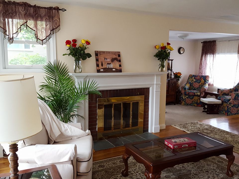 A comfortable living space features a classic fireplace with a brass screen, surrounded by decorative floral arrangements. Two armchairs with colorful upholstery are positioned near a wooden coffee table, while an area rug adds warmth to the hardwood floor.