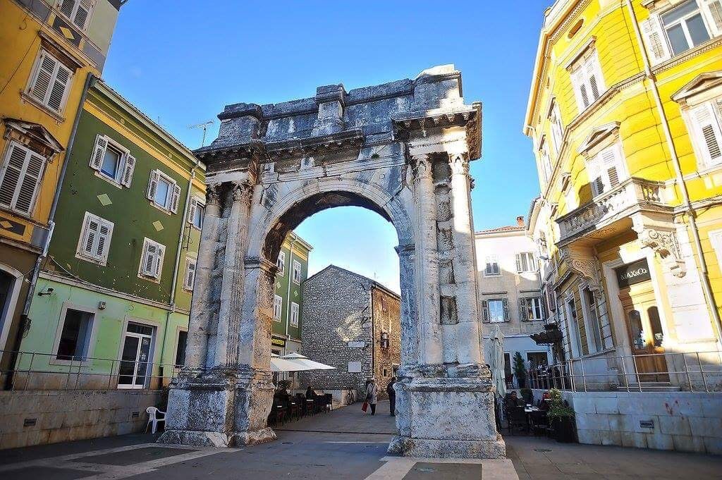 An ancient stone arch stands prominently in a bustling square, surrounded by colorful buildings with various window styles. Tables and chairs are arranged nearby, inviting visitors to relax and enjoy the lively atmosphere of the area.