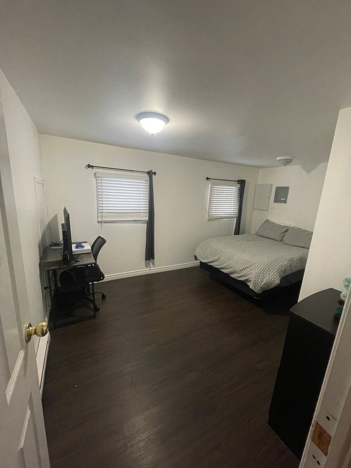 A well-lit bedroom features a spacious bed with a neutral patterned comforter. Two small windows are dressed with blackout curtains, allowing managed natural light. A desk with a chair is positioned against the wall, complemented by a dark wood dresser nearby.