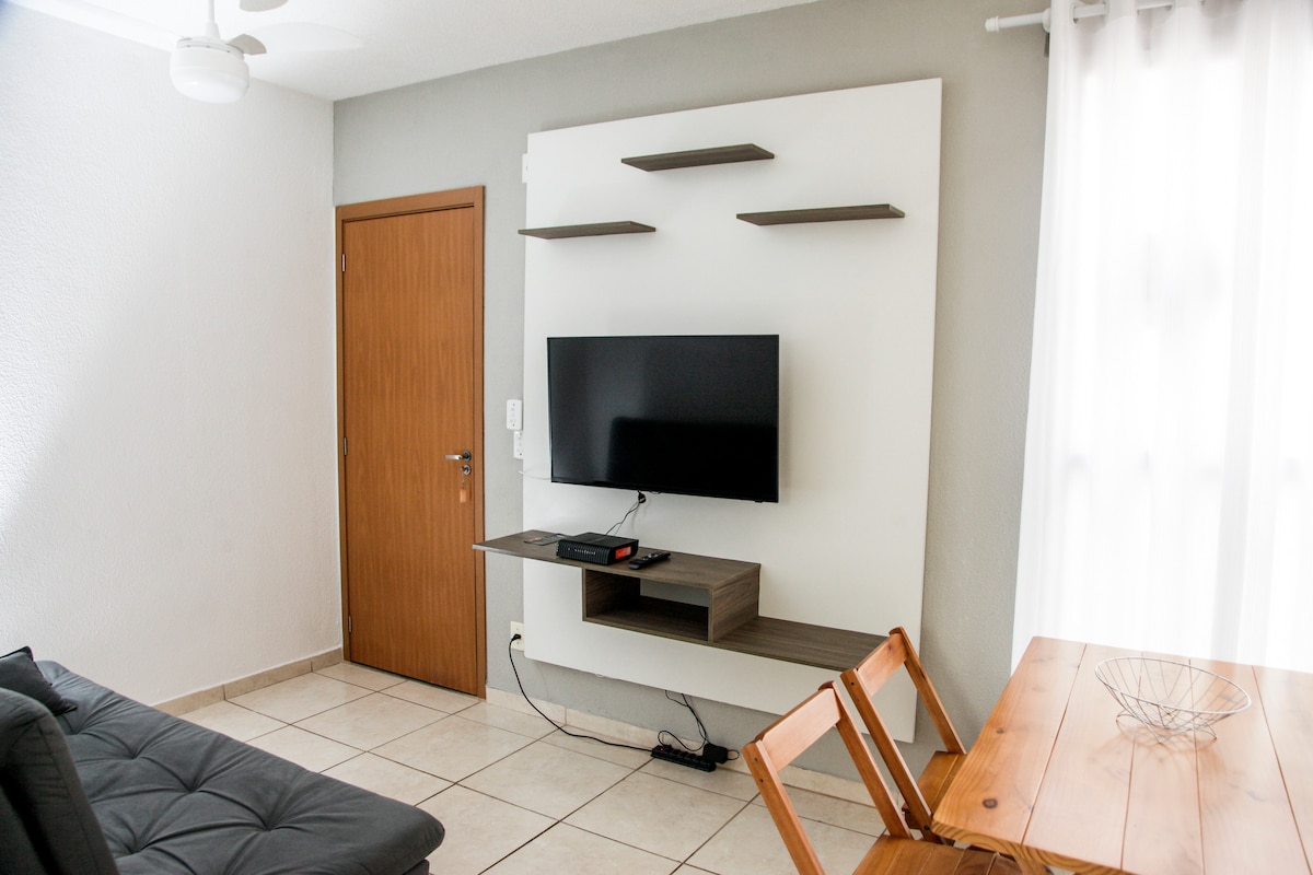 A modern living area is highlighted by a large flat-screen television mounted on a feature wall. A wooden table with two matching chairs is positioned nearby, while natural light enters through sheer curtains, enhancing the spacious feel of the room.