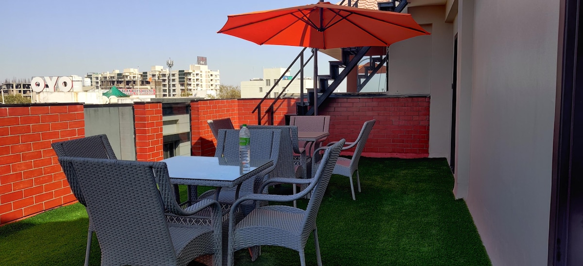 An outdoor terrace is equipped with a white table surrounded by six gray wicker chairs. An orange umbrella provides shade, while artificial grass covers the floor. The background displays city buildings, visible under clear skies.