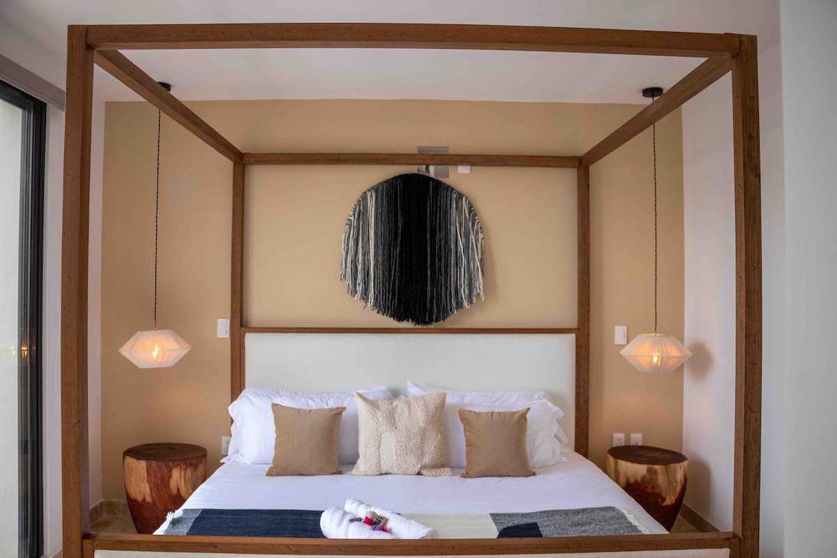 A spacious bed is framed by a wooden four-poster structure, complemented by two bedside pendant lights. Neutral-colored cushions add comfort, while a wall art piece creates visual interest. The room features smooth walls and wooden side tables, contributing to a relaxing atmosphere.