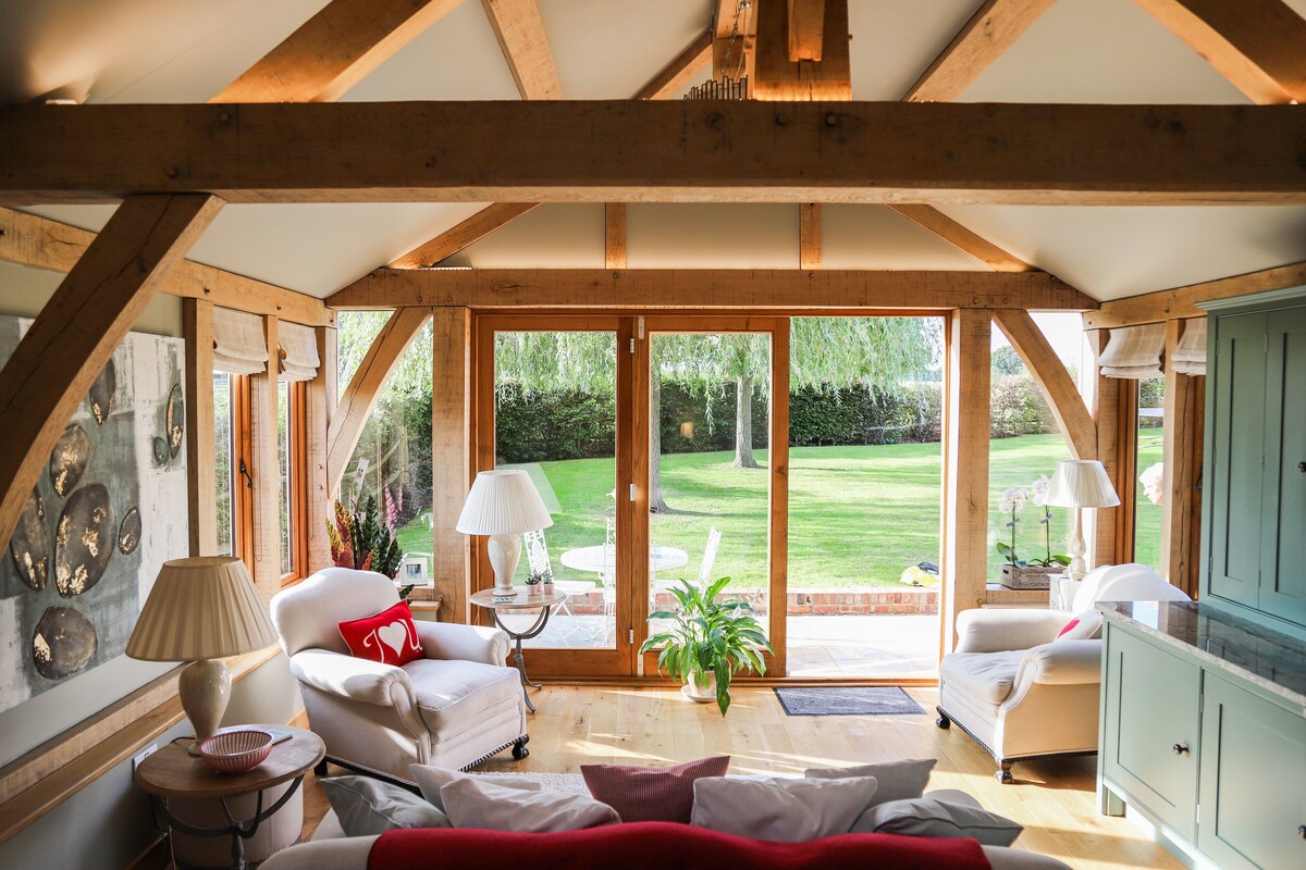 A cozy living area features exposed wooden beams and bi-fold doors that open to a private patio and a well-maintained lawn. A pair of comfortable chairs is positioned near a lamp, with a subtle arrangement of decorative elements on the side table.