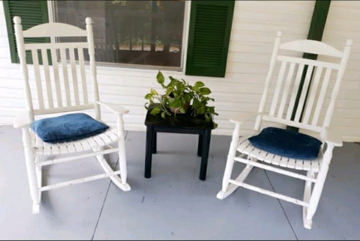 Two white rocking chairs with blue cushions are positioned beside a small black table on a covered porch. A potted plant sits atop the table, adding a touch of greenery to the inviting seating area.