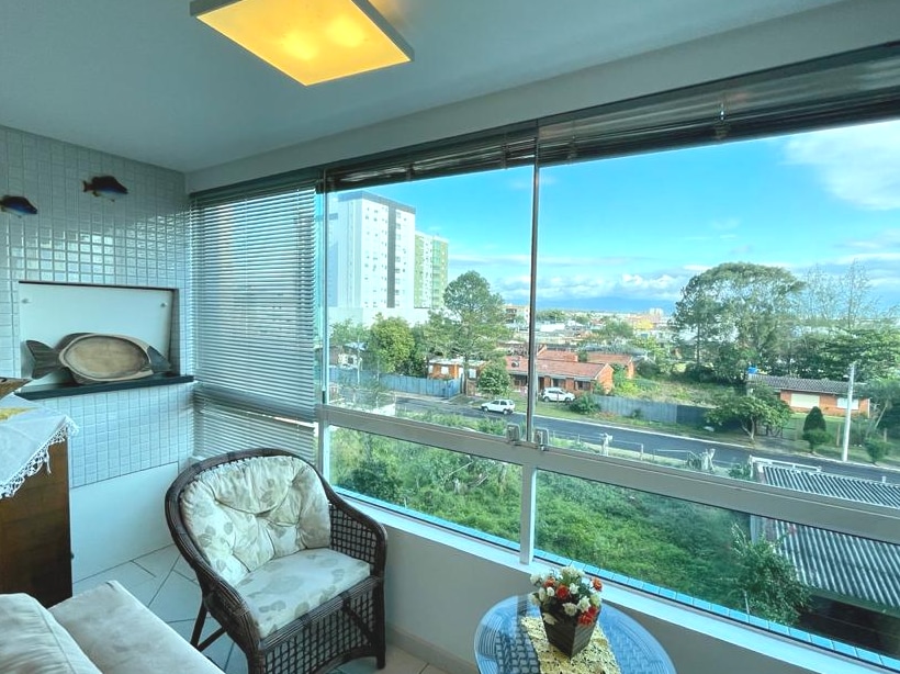 A well-lit seating area features two comfortable chairs positioned beside a glass table. Large windows allow for natural light and offer a view of the surrounding greenery and distant ocean. A decorative plant sits on the table, enhancing the serene atmosphere.