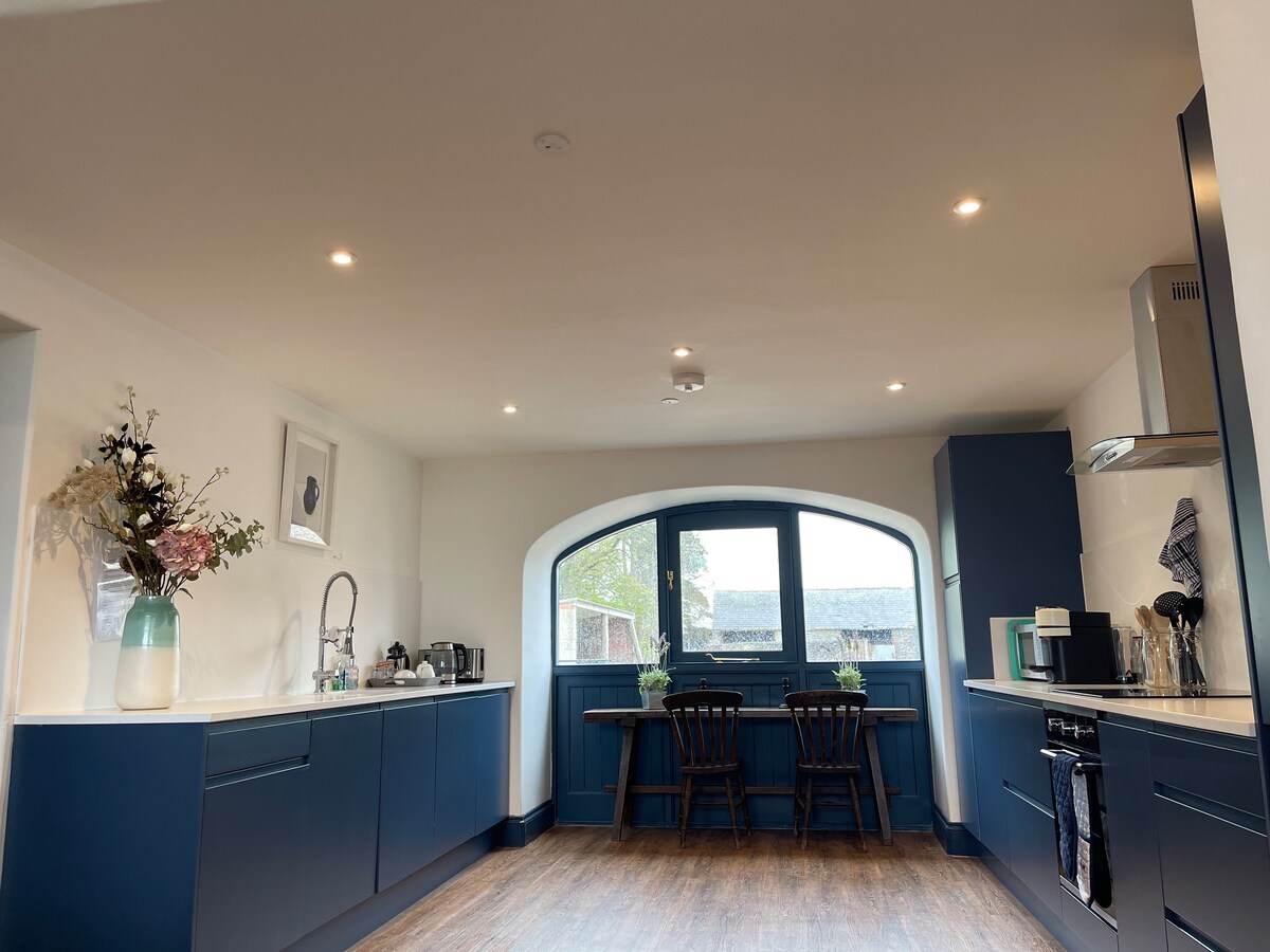 A spacious kitchen features modern blue cabinetry and a light countertop. Natural light pours through an arched window, illuminating the seating area with a wooden table and chairs. Stylish decor includes a vase with flowers, creating a functional and inviting cooking space.