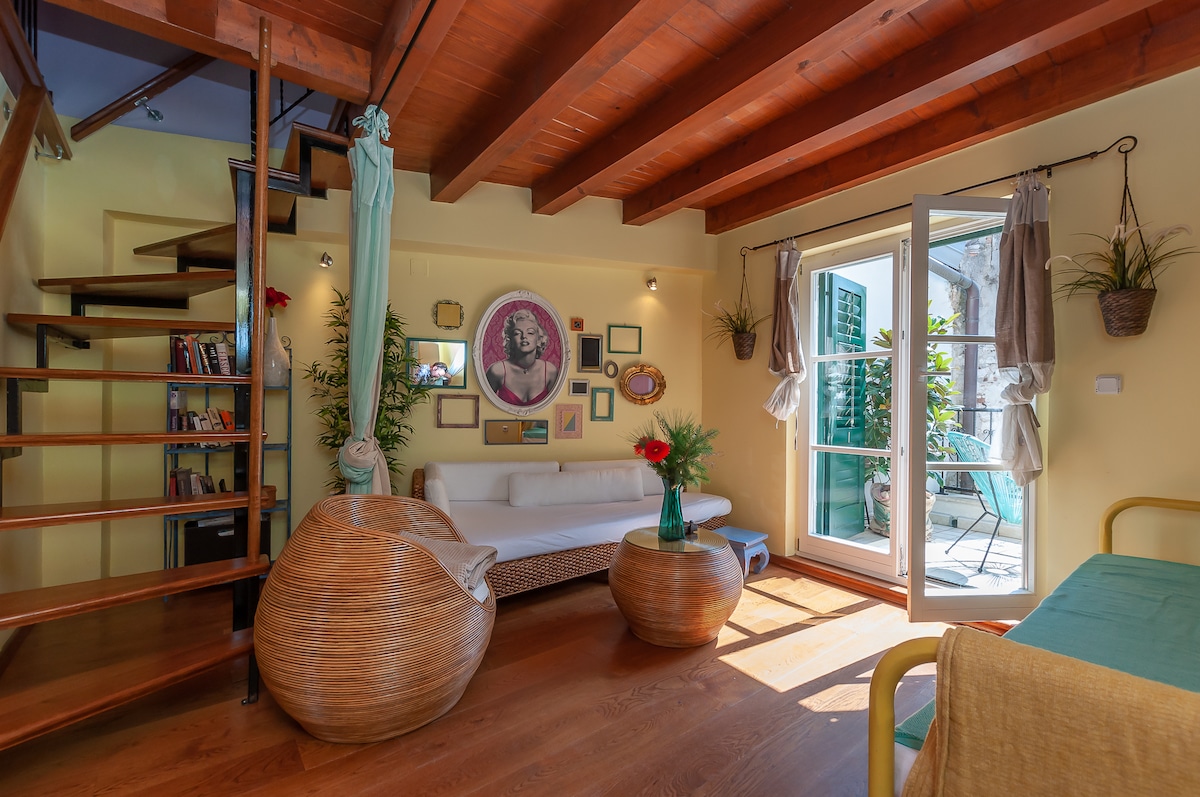A living area features wooden beams and a light-toned wall, contributing to a cozy ambiance. A white sofa is positioned along the wall, while potted plants and decorative elements are placed throughout. Large doors open to a balcony, providing natural light and outdoor access.