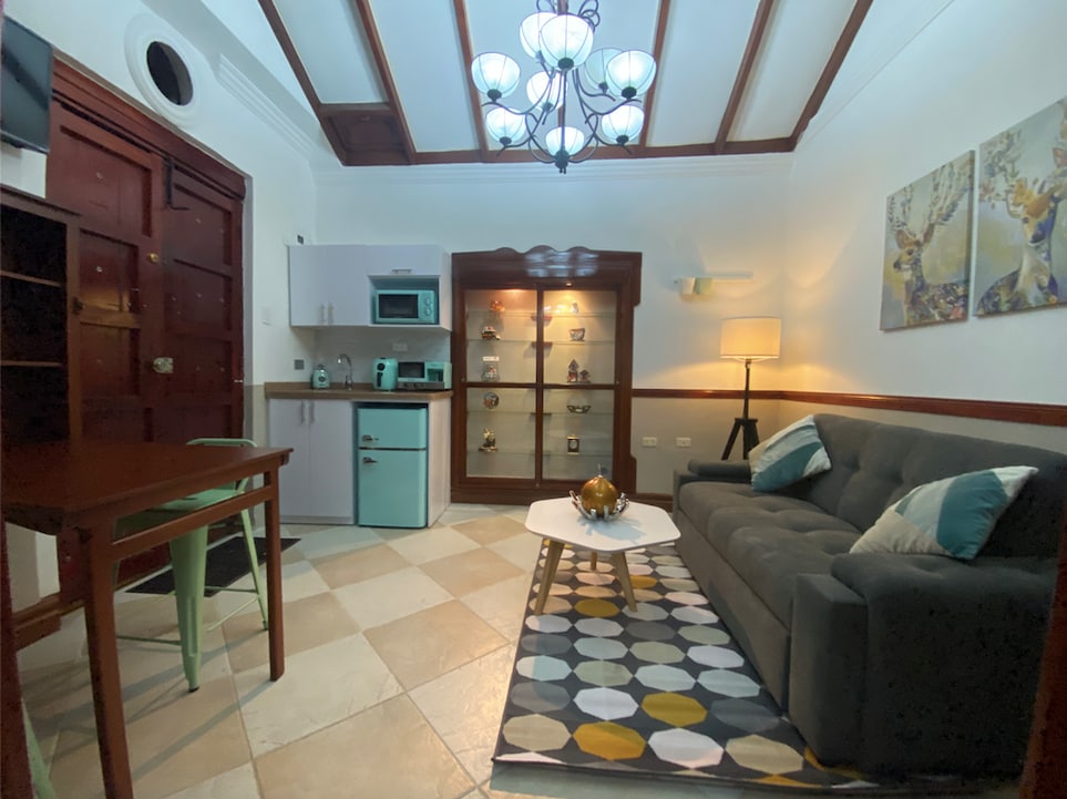 A living area is displayed featuring a dark gray sofa and a light-colored coffee table. A dining table with two chairs is positioned nearby. Wall shelves exhibit decorative items, and a modern chandelier illuminates the space. The floor is tiled in a light-colored pattern.