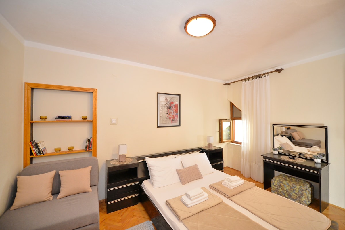 A well-organized bedroom is presented with two spacious beds, complemented by a cozy seating area. Natural light enters through the window, while a mirror and a small bookshelf enhance the room's functionality. Soft, neutral tones contribute to a calm environment.