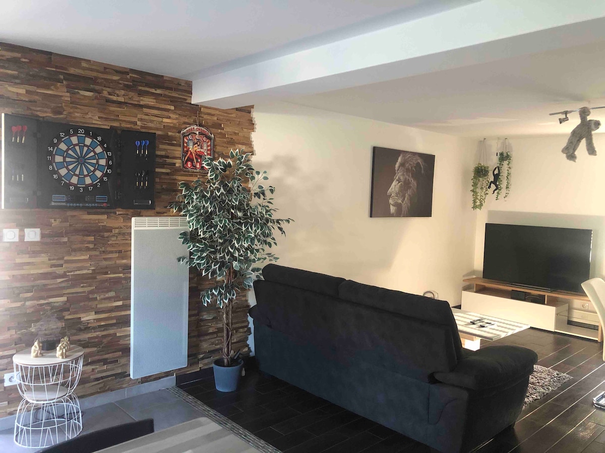 A modern living area is presented with a dark couch facing a wall-mounted television and a mounted electronic dartboard above a textured wood accent wall. A decorative plant is positioned nearby, enhancing the space's ambiance.